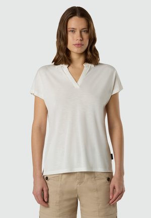 Donna con capelli castani di media lunghezza, che indossa una maglietta bianca a maniche corte con scollo a V e pantaloncini cargo beige, in piedi davanti a uno sfondo semplice.