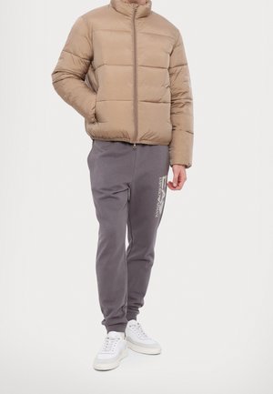 Mand iført en beige dunjakke, grå joggingbukser med Emporio Armani-logo og hvide sneakers, stående mod en ensfarvet baggrund.