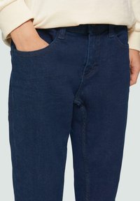 Donkerblauwe denim jeans met een rechte pijpdesign, voorzien van voorzakken en zichtbare stikseldetails. De stof heeft een gladde textuur.