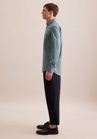 Lichtblauw overhemd met knoopsluiting en een zak, gecombineerd met donker marineblauwe broek en zwarte nette schoenen, van opzij weergegeven tegen een neutrale achtergrond.
