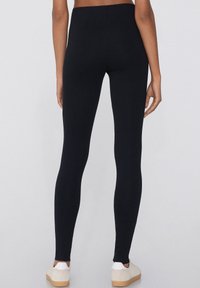 Leggings noirs avec une texture lisse, taille haute et coupe ajustée, associés à des chaussures blanches et beiges sur un fond uni.