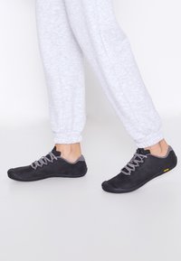 Scarpe nere con accenti grigi, realizzate in materiale simile alla suede. Presentano suole in gomma e lacci. Indossate con pantaloni da jogging grigi chiari.