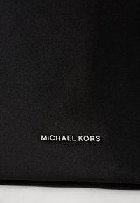 Sac en tissu noir avec une surface texturée, arborant un logo argenté "MICHAEL KORS" bien en vue. Design simple sans accents supplémentaires.