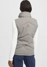 Padded sivi i crni kvadratasti prsluk s visokom ovratnikom, s teksturiranom površinom i šivenim detaljima, nošen preko bijele majice dugih rukava.