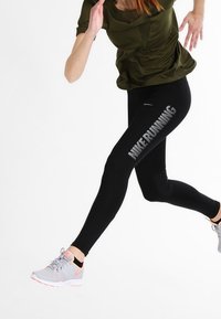 Osoba w trakcie biegu, ubrana w czarne legginsy z napisem "NIKE RUNNING", oliwkową koszulkę i szare tenisówki Nike z pomarańczowymi akcentami, na białym tle.