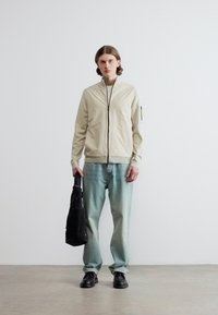 Νεαρό άτομο με μαλλιά μέχρι τους ώμους φοράει μπεζ μπουφάν με φερμουάρ, ανοιχτό μπλε τζιν, μαύρα παπούτσια και κρατάει μια μαύρη τσάντα σε απλό φόντο.