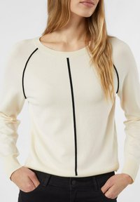 Frau trägt einen cremefarbenen Pullover mit schwarzen vertikalen und geschwungenen Linien, Hände in den Taschen einer schwarzen Hose, vor einfachem weißem Hintergrund.