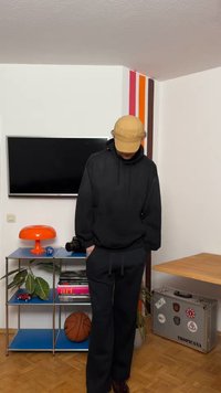 Isik, kes kannab musta kapuutsiga pusa ja pükse, ning beežikat mütsi, vaatab allapoole toas, kus on oranž lamp, korvpall, teler ja hõbedane kleebistega kohver.