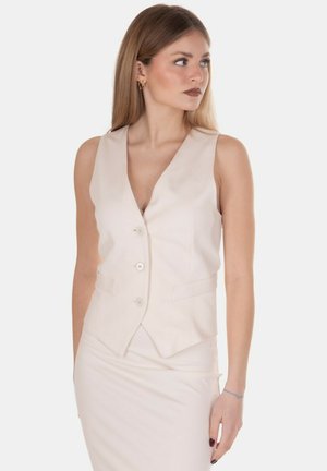 Giovane donna che guarda a destra, indossa un gilet crema senza maniche con bottoni e gonna abbinata su sfondo bianco uniforme.