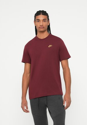 Uomo con capelli intrecciati che indossa una maglietta Nike color bordeaux e pantaloni di tuta Nike grigio scuro, in piedi su uno sfondo bianco.