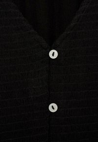 Black textured fabric with a v-neckline, featuring two white buttons near the center. The material has subtle horizontal ridges.