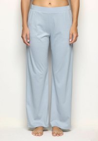 Mujer vestida con pantalones de pijama sueltos de color azul claro, de pie descalza frente a un fondo liso, mostrando la parte media del cuerpo hasta los pies.