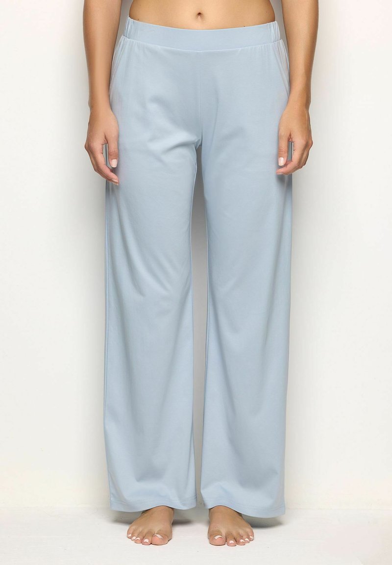 Mujer vestida con pantalones de pijama sueltos de color azul claro, de pie descalza frente a un fondo liso, mostrando la parte media del cuerpo hasta los pies.