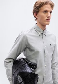 Γκρι πουκάμισο με κουμπιά και υφή, με μικρό μαύρο λογότυπο, συνδυασμένο με μαύρη υφαντή τσάντα duffel.