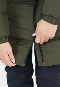 Chaqueta de plumas verde oliva con apertura con cremallera, presenta puños elásticos. Llevada sobre pantalones oscuros, con las manos agarrando la cremallera.