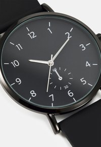 Montre analogique noire au design minimaliste. Elle dispose d'un cadran noir, d'aiguilles des heures et des minutes en argent, et d'un petit sous-cadran pour les secondes.