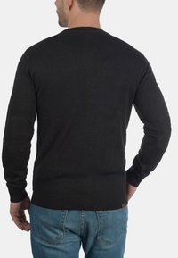Mörkgrå stickad tröja med rund halsringning, ribbade mudd och nederkant, försedd med armbågsfickor. Bärs över ljusblå jeans.