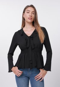Blouse noire à manches longues, col à volants et détail de nœud au centre. Taille smockée et ourlet évasé pour une silhouette fluide.