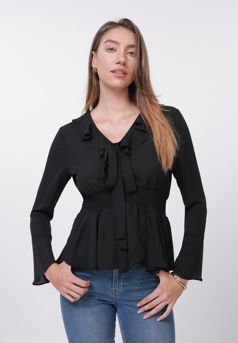Blouse noire à manches longues, col à volants et détail de nœud au centre. Taille smockée et ourlet évasé pour une silhouette fluide.
