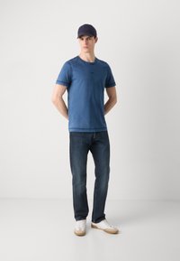 Μπλε μπλούζα με λαιμόκοψη και κοντά μανίκια, σε συνδυασμό με σκούρους τζιν παντελόνι και άσπρα αθλητικά παπούτσια με μπεζ λεπτομέρειες. Ένα ναυτικό καπέλο ολοκληρώνει την εμφάνιση.