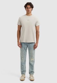 Lichtgrijze, korte mouwen t-shirt met een minimalistisch zwart logo op de borst, gecombineerd met lichtblauwe versleten spijkerbroek en lichtbruine schoenen.