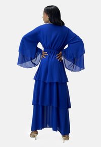Lang blå kjole med lagdelte chiffonlag, brede klokkeærmer, markeret talje og ankellang længde. Har en glat, flydende tekstur.