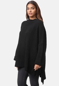 Maglione nero oversize in maglia con collo alto, texture a coste, maniche lunghe e orlo arcuato. Indossato sopra dei jeans neri.