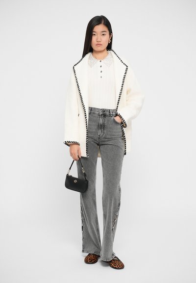 Veste en mélange de laine blanche avec des détails noirs, haut côtelé crème, jean en denim gris taille haute avec fentes sur les côtés, et chaussures slip-on imprimé léopard. Sac à main noir.