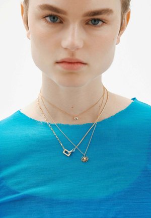 Collares de oro en estilos en capas, con un pequeño colgante geométrico, un colgante de cristal redondo y un motivo de ojo. Llevados sobre una blusa azul de punto acanalado.