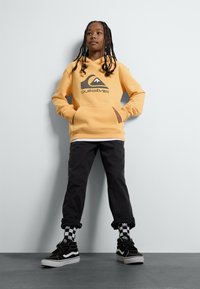 Gul huvtröja med en svart logotyp, svarta byxor och svarta sneakers med vita sulor. Har rutiga strumpor och en avslappnad passform.