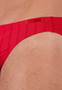 Czerwony strój kąpielowy w formie slipów z poziomymi paskami, miękką teksturą oraz marki elastycznym paskiem w talii z widoczną metką z logo.