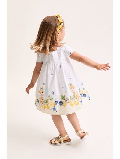 Kisgyermek lány fehér ruhában, amelyen kék csillagok és rajzfilmfigura található, virágmintás szandált és fejpántot visel, és pörög.