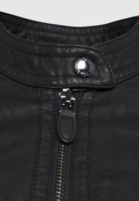 Veste en cuir noir avec un col montant, dotée d'un bouton-pression en argent, d'une fermeture éclair apparente et d'une surface douce et texturée.