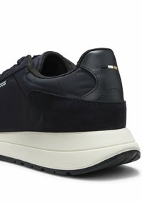 Gros plan du talon d'une basket noire avec des matériaux mixtes, une semelle blanche épaisse et un petit détail cousu noir, blanc et beige sur la languette du talon.