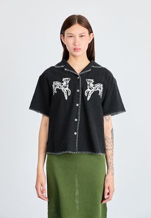Black short-sleeve shirt with white embroidered deer motifs, button-up front, contrast stitching, and a relaxed fit, worn over a green textured skirt.