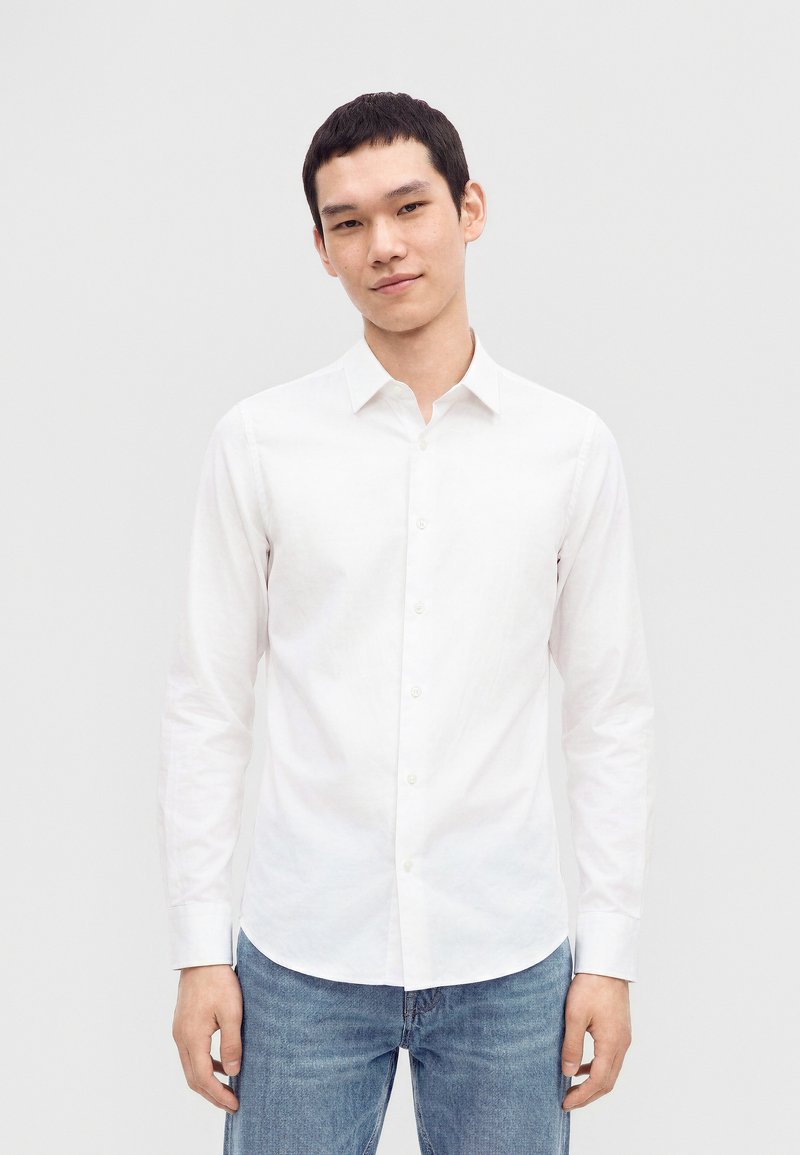 Jeune homme aux cheveux courts noirs portant une chemise blanche boutonnée et un jean bleu, debout devant un fond clair uni.