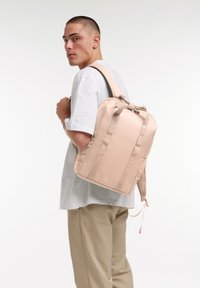 Mochila de color rosa claro, hecha de tela con una textura suave, que cuenta con dos correas de hombro resistentes y un asa superior. Diseño sencillo con acentos mínimos.