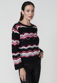 Pull noir avec des motifs zigzag multicolores en rouge, gris et crème. Il a des manches longues et un col rond. Tissu en maille texturée.