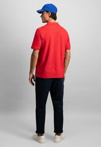 Rød polo skjorte med korte ermer, iført mørke bukser og hvite sneakers. Blå caps og enkel design. Enfarget bakgrunn.