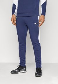 Marineblauwe sport joggers met witte zijstrepen en een logo op de linker bovenbeen; gemaakt van een gladde stof en een slim fit ontwerp. Zwarte sportschoenen.