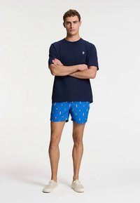 Lagana tamnoplava majica kratkih rukava, plave kupaće gaće s šarenim uzorcima i bijele tenisice. Ležerna odjeća s opuštenim krojem i laganim materijalom.