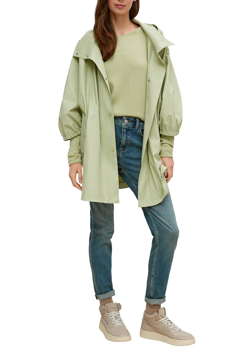 Jasnozielona wodoodporna kurtka z przerośniętymi, bufiastymi rękawami, w połączeniu z jasnozielonym swetrem, niebieskimi dżinsami i beżowymi trampkami za kostkę.