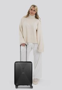 Valigia rigida nera con texture scanalata, manico retrattile e quattro ruote, posizionata accanto a una persona in un ampio maglione chiaro e pantaloni bianchi.