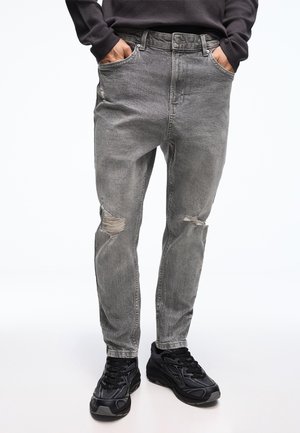 Personne portant un jean gris usé et des baskets noires, les mains dans les poches, debout sur un fond blanc.