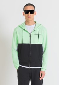 Felpa con zip di colore verde chiaro e nero con cappuccio, dotata di zip argentata e lacci neri. Indossata sopra una t-shirt bianca.