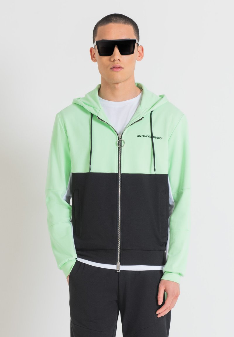 Felpa con zip di colore verde chiaro e nero con cappuccio, dotata di zip argentata e lacci neri. Indossata sopra una t-shirt bianca.