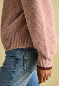 Rosa, strukturierter Pullover mit geripptem Saum und Ärmeln; dunkellila Akzent an den Ärmeln. Kombiniert mit blauen Jeans. Weiches, grob gestricktes Material.