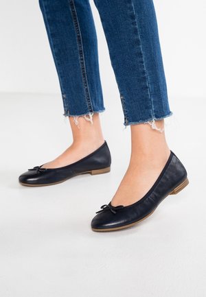 Fødder iført sorte læder-ballerinasko med små sløjfer, stylet med blå cropped jeans med frynset kant på hvidt gulv.