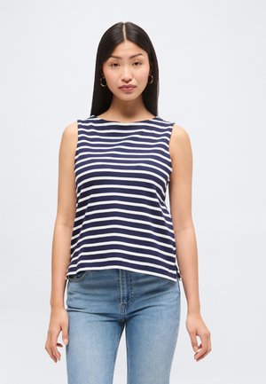 Kobieta o długich ciemnych włosach, nosząca złote koła kolczyków, granatowo-biały, pasiasty top bez rękawów oraz jasnoniebieskie dżinsy, stojąca na jednolitym tle.