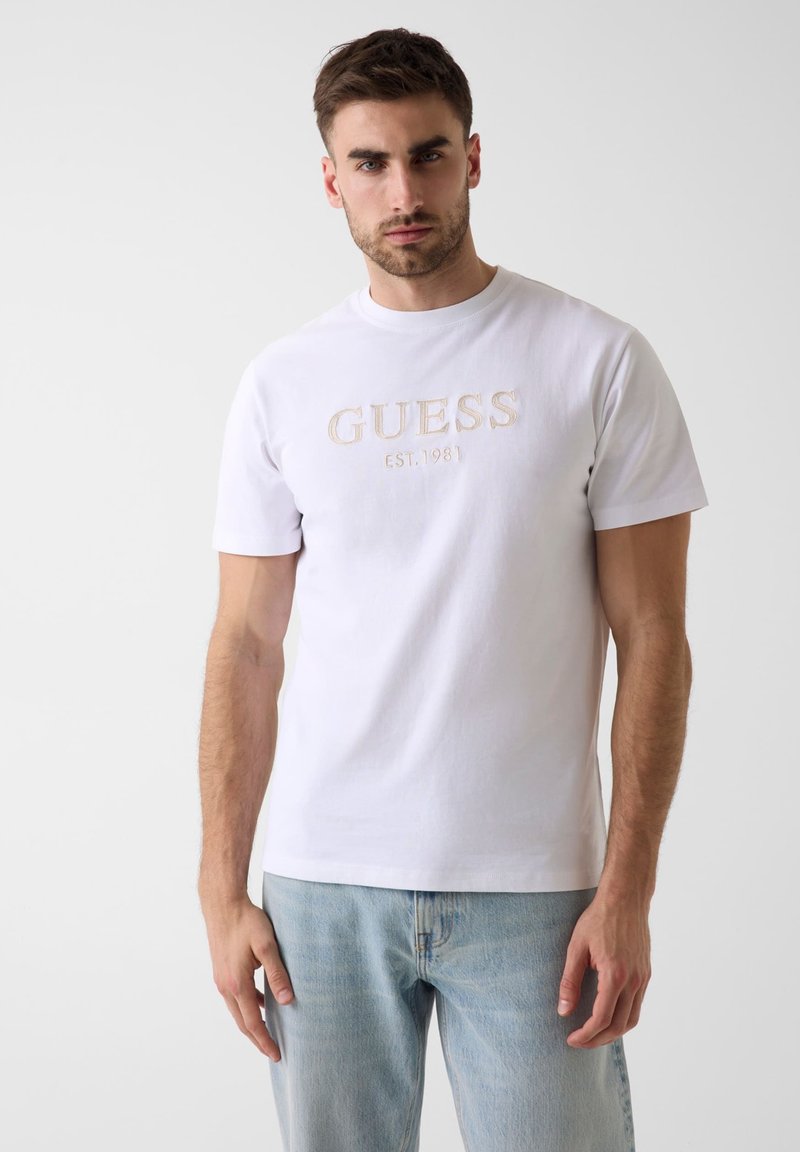 Homme portant un t-shirt blanc à manches courtes avec le texte « GUESS EST. 1981 » et un jean bleu clair, debout devant un fond uni gris clair.